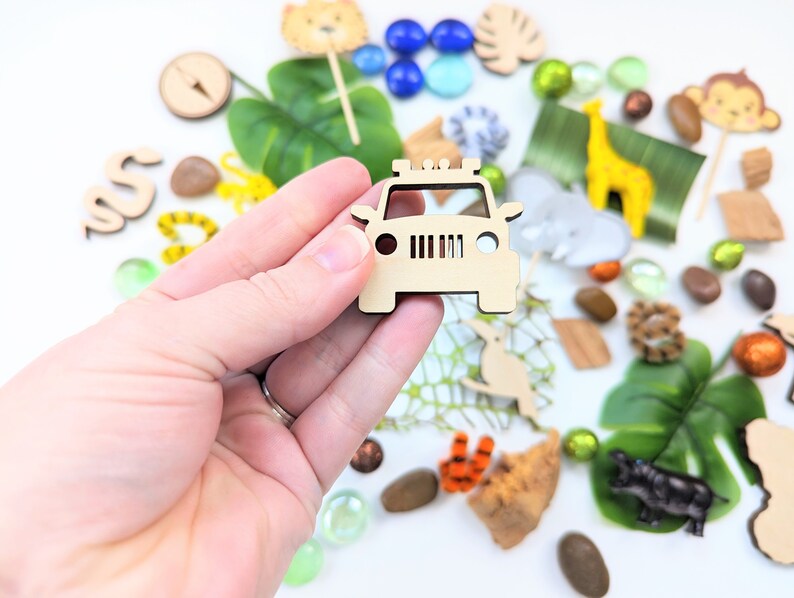 May include: Wooden toy jeep cutout, a part of a larger set of safari-themed toys. The jeep is light brown and has a simple design with a grill, headlights, and a roof rack. The jeep is surrounded by other safari-themed toys, including a giraffe, a monkey, a snake, and a compass.