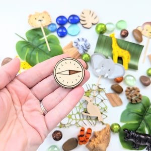 May include: A wooden compass with a brown background and a black arrow pointing north. The compass is surrounded by a variety of jungle-themed toys and decorations, including leaves, animals, and beads.