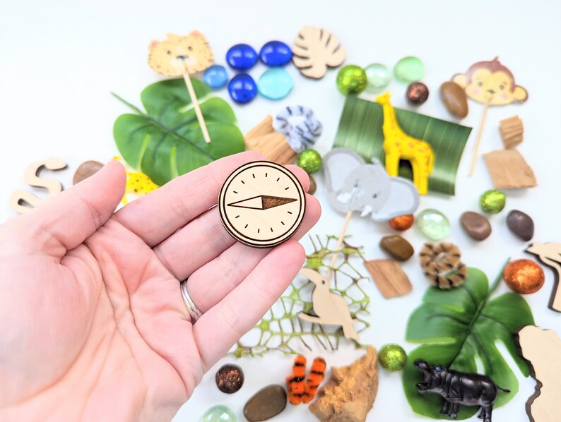 May include: A wooden compass with a brown background and a black arrow pointing north. The compass is surrounded by a variety of jungle-themed toys and decorations, including leaves, animals, and beads.