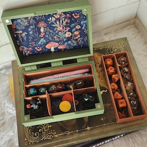 May include: An open sage green wooden box with a floral mushroom print lining on the lid, containing a pink pen, multi-colored polyhedral dice, and small game tokens. Beside it sits a separate rectangular orange tray filled with orange and clear dice.