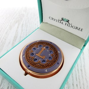 May include: A round, purple compact with a gold filigree design and a crystal 'L' in the center. The compact is in a teal box with the words 'Crystal Filigree' on the lid.