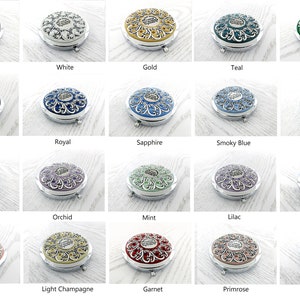 May include: A collection of 24 compact mirrors with ornate silver frames and various colored enamel inlays. The mirrors are arranged in four rows of six, with each row featuring a different color scheme.