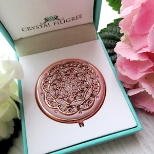 May include: A rose gold compact mirror with a pink and white crystal filigree design. The mirror is in a turquoise box with the words "Crystal Filigree" on the lid.
