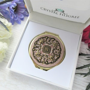 May include: A pink compact mirror with a gold filigree design and a crystal 'N' in the center. The mirror is in a white box with the words "Crystal Filigree" printed on the top.