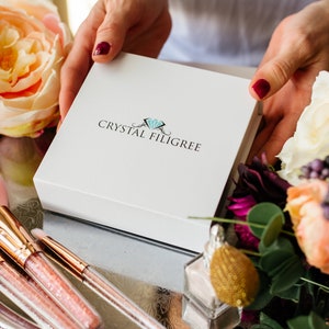 May include: A white box with the text "CRYSTAL FILLIGREE" and a blue diamond logo. The box is being held by hands with red nail polish. The box is surrounded by pink and white flowers.