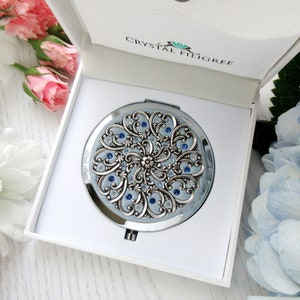 Op de afbeelding: Een zilveren compact spiegel met een blauw en zilver filigraanontwerp en blauwe kristallen accenten. De spiegel zit in een witte doos met de woorden "Crystal Filigree" erop gedrukt.