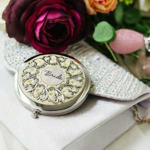 Puede incluir: Un espejo compacto plateado con la palabra "Bride" en escritura, apoyado en un clutch blanco. El espejo tiene un dise&ntilde;o floral ornamentado. Un ramo de flores y un frasco de perfume est&aacute;n en el fondo.