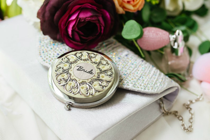 Puede incluir: Un espejo compacto plateado con la palabra "Bride" en escritura, apoyado en un clutch blanco. El espejo tiene un dise&ntilde;o floral ornamentado. Un ramo de flores y un frasco de perfume est&aacute;n en el fondo.
