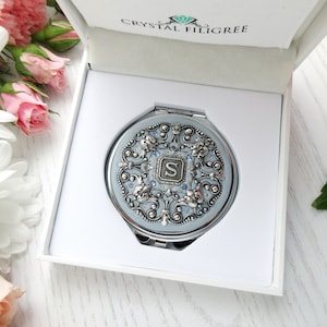 May include: A silver compact mirror with a blue background and a silver filigree design. The mirror has a crystal embellishment and the letter "S" in the center.