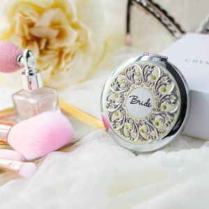 Puede incluir: Un espejo compacto plateado con un dise&ntilde;o ornamentado y la palabra "Bride" en el centro. El espejo est&aacute; rodeado de brochas de maquillaje, un frasco de perfume y una caja blanca. El tema general es nupcial y belleza.