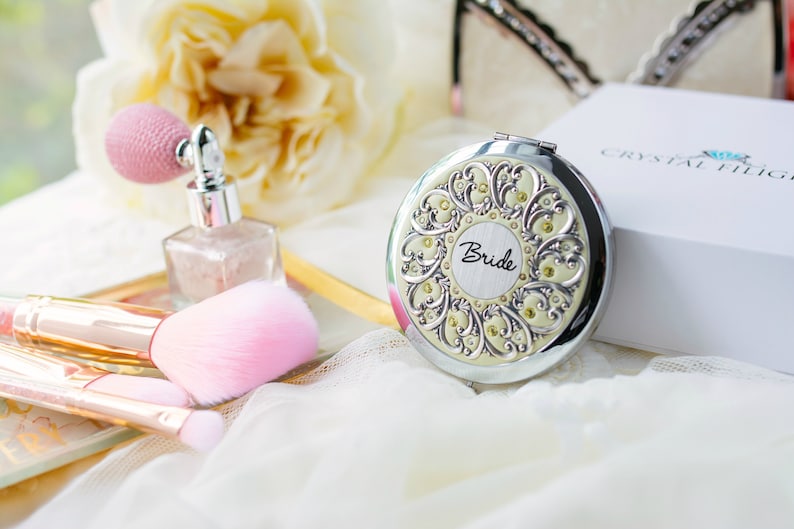 Puede incluir: Un espejo compacto plateado con un dise&ntilde;o ornamentado y la palabra "Bride" en el centro. El espejo est&aacute; rodeado de brochas de maquillaje, un frasco de perfume y una caja blanca. El tema general es nupcial y belleza.