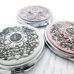 Puede incluir: Tres espejos compactos decorativos en blanco, lavanda y rosa. Cada compacto presenta un diseño plateado ornamentado con una inicial central, rodeado de acentos brillantes. Los compactos tienen un marco de metal plateado.