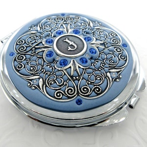 May include: A silver compact mirror with a blue enamel background. The mirror has a decorative design with a silver filigree and blue rhinestones. The centre of the mirror features a silver letter "J" surrounded by rhinestones.