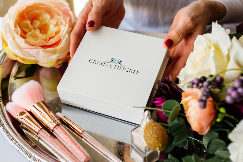 Puede incluir: Una caja blanca con el texto "CRYSTAL FILIGREE" es sostenida por manos con esmalte de u&ntilde;as rojo, rodeada de brochas de maquillaje, flores y un frasco de perfume en una bandeja plateada. La caja es el punto focal de la imagen.