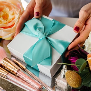 May include: A white gift box with a turquoise ribbon tied in a bow. The box is surrounded by makeup brushes and flowers.