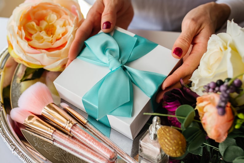 May include: A white gift box with a turquoise ribbon tied in a bow. The box is surrounded by makeup brushes and flowers.