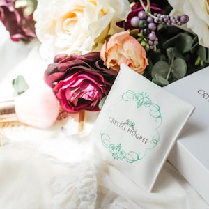 Puede incluir: Imagen de una caja blanca y una peque&ntilde;a bolsa, ambas con la marca "Crystal Filigree" en una escritura elegante. La bolsa tiene un dise&ntilde;o de filigrana turquesa. La escena se complementa con un ramo de rosas y otras flores, y un frasco de perfume.