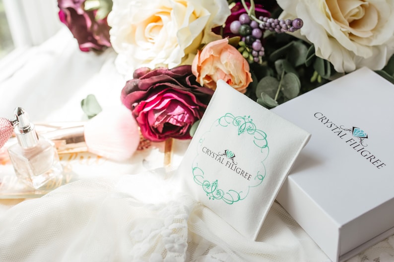 Puede incluir: Imagen de una caja blanca y una peque&ntilde;a bolsa, ambas con la marca "Crystal Filigree" en una escritura elegante. La bolsa tiene un dise&ntilde;o de filigrana turquesa. La escena se complementa con un ramo de rosas y otras flores, y un frasco de perfume.