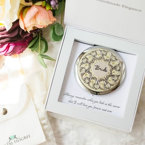 Puede incluir: Un espejo compacto plateado con un dise&ntilde;o ornamentado y la palabra "Bride" en el centro, presentado en una caja blanca. La caja contiene una tarjeta con un mensaje de amor. Las palabras "Handmade Elegance" est&aacute;n en la parte superior de la caja.