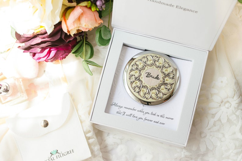 Puede incluir: Un espejo compacto plateado con un dise&ntilde;o ornamentado y la palabra "Bride" en el centro, presentado en una caja blanca. La caja contiene una tarjeta con un mensaje de amor. Las palabras "Handmade Elegance" est&aacute;n en la parte superior de la caja.