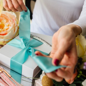 May include: A white gift box with a turquoise ribbon tied in a bow. The ribbon is being untied by a hand.