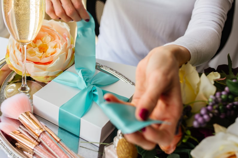 May include: A white gift box with a turquoise ribbon tied in a bow. The ribbon is being untied by a hand.