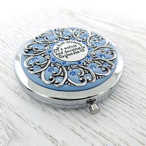 May include: A silver compact mirror with a blue background and a silver filigree design. The mirror has a quote that reads "The love between a mother and daughter is forever".