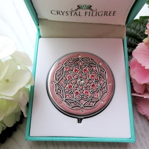May include: A pink compact mirror with a silver filigree design and crystal accents. The mirror is in a turquoise box with the words "Crystal Filigree" printed on the top.