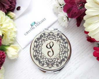May include: A silver compact mirror with a white background and a silver filigree design. The center of the mirror features a white circle with a gold letter "S" in the center. The mirror is surrounded by a white background with the text "Crystal Filigree" printed on it.