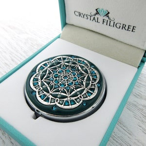 Pode incluir: Um espelho compacto turquesa com um design de filigrana prateada e cristais azuis e brancos. O espelho está em uma caixa turquesa com as palavras "Crystal Filigree" na tampa.