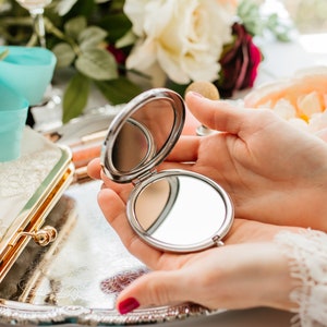 May include: A silver compact mirror with a round, reflective surface. The mirror is open and being held by a person's hands.