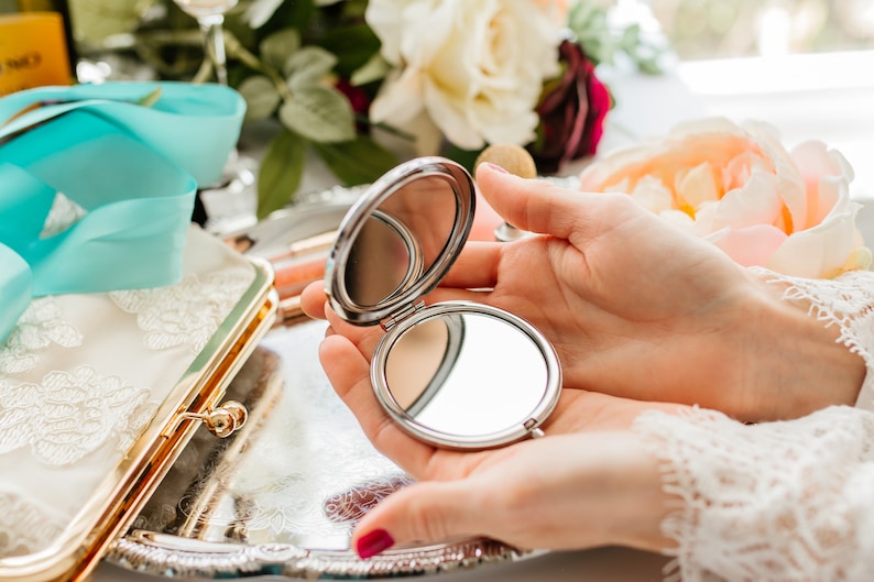 May include: A silver compact mirror with a round, reflective surface. The mirror is open and being held by a person's hands.