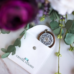 Puede incluir: Un colgante redondo con un marco plateado y un diseño decorativo de cristales transparentes y una letra "B" marrón se muestra en una bolsa blanca. La bolsa se cierra con un broche plateado. La marca "CRYSTAL FILIGREE" está impresa.