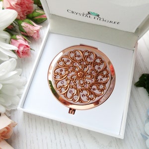 Op de afbeelding: Een compact spiegel in rosé goud met een roze en gouden filigraan ontwerp en heldere kristallen. De spiegel zit in een witte doos met de tekst "Crystal Filigree".