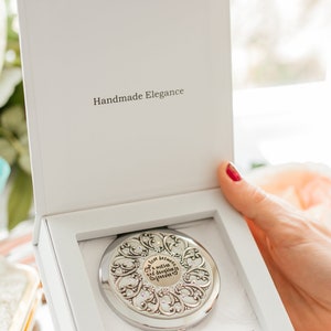 May include: A silver compact mirror with a decorative design and a quote that reads "The love between a mother and daughter is forever". The mirror is in a white box with the words "Handmade Elegance" printed on the lid.