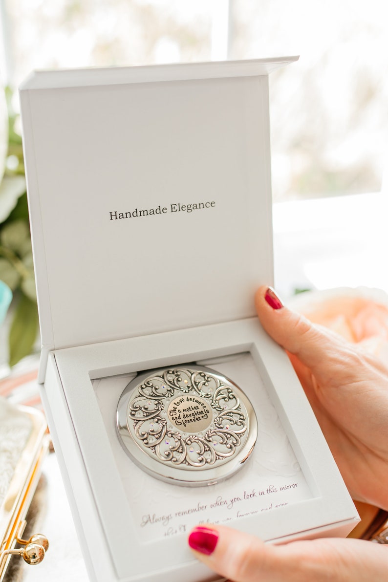 May include: A silver compact mirror with a decorative design and a quote that reads "The love between a mother and daughter is forever". The mirror is in a white box with the words "Handmade Elegance" printed on the lid.