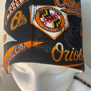 May include: Black scrub hat with an orange and white Baltimore Orioles baseball team print. The hat has a tie string.