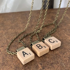 Può includere: Tre collane a catena dorata con ciondoli a forma di tessere di lettere in legno. Le tessere compongono la parola "A", "B" e "C".
