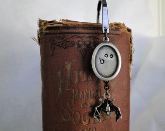 Puede incluir: Marcapáginas de plata con un amuleto de murciélago y una etiqueta redonda que dice "boo" con dos puntos para los ojos. El marcapáginas está unido a un libro con una cubierta marrón.