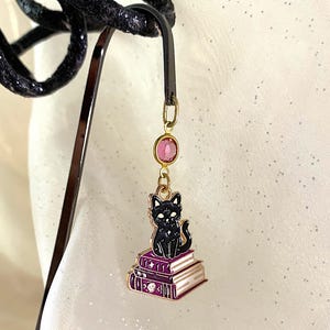 May include: A gold bookmark with a black cat sitting on top of three purple books. The cat has white eyes and a pink gem hangs from the top of the bookmark.