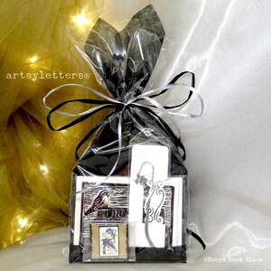 May include: A gift package wrapped in black and clear plastic with a white ribbon. The package contains a bookmark with a bird design, a card with a bird design, and a small silver charm. The package is labeled "artsylettters®".