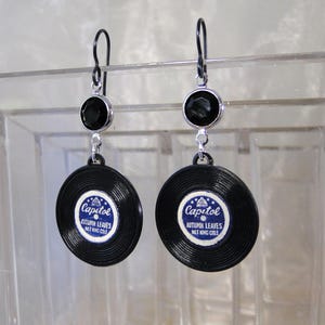 May include: A pair of black and silver earrings featuring miniature vinyl records with the text "Capitol Autumn Leaves Nat King Cole" printed on them. Each earring has a black gemstone at the top.