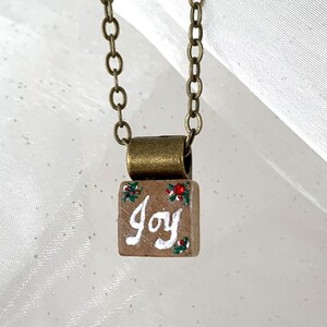May include: A bronze necklace featuring a small, rectangular pendant. The pendant is painted brown with the word "Joy" in white script, decorated with red berries and green leaves. The chain is also bronze.