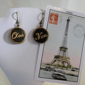 Op de afbeelding: Een paar gouden oorbellen met zwarte ronde hangers. De hangers hebben gouden letters met de woorden "Oui" en "Non".