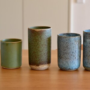 Op de afbeelding: Vier keramische bekers in verschillende maten en kleuren staan op een houten oppervlak. De kleuren zijn groen, blauw en een gespikkeld blauwgrijs. De bekers hebben een cilindrische vorm met een licht afgeronde basis.