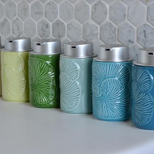 May include: A row of ceramic soap dispensers in various colors, including white, yellow-green, green, teal, and blue. Each dispenser has a silver top and a textured, fan-like pattern. They are arranged on a white surface against a patterned tile backdrop.