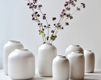 Può includere: Una collezione di sei vasi in ceramica bianca opaca con corpi arrotondati e aperture strette. Un vaso centrale contiene un rametto di erbe con fiori viola. I recipienti variano in altezza e larghezza, presentando superfici lisce e uniformi.