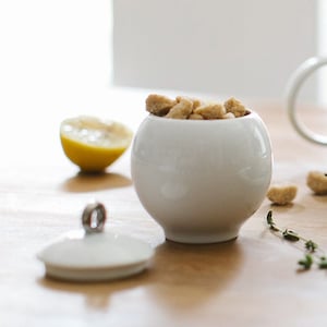 以下が含まれることがあります： 蓋付きの白いセラミック製の砂糖入れで、茶色の角砂糖が入っています。砂糖入れは、レモンのくさび形とタイムの枝が置かれた木製のテーブルの上にあります。