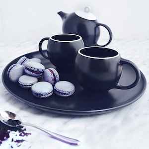 Puede incluir: Una bandeja negra con dos tazas de té negras y una tetera negra. La bandeja también tiene una pila de macarons de color lavanda.