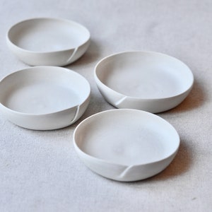 May include: Four white ceramic bowls with a unique, slightly uneven rim. The bowls are arranged in a staggered pattern on a light gray fabric surface.